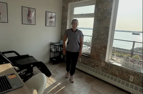 This is a brightly lit photo of a woman in grey professional scrubs, smiling as she stands in our collaboration clinic room. To her left is a medical examination chair and anatomical charts on the wall. The room features a large window with a marbled frame that offers a scenic view of a wide body of water, with what looks like train tracks running along the shoreline below.