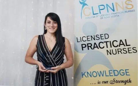 A smiling woman in a black and white striped dress holds an award while standing next to a banner for the College of Licensed Practical Nurses of Nova Scotia (CLPNNS). The banner reads "LICENSED PRACTICAL NURSES" and "KNOWLEDGE is our Strength." The background features soft, glowing lights, suggesting a formal recognition event.
