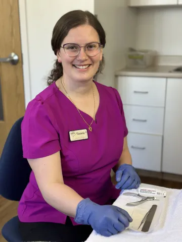 A smiling foot care nurse wearing a magenta scrub top and blue gloves sits at a treatment station. She has a name tag that reads “Victoria – LPN Nursing” and is positioned beside a neatly arranged set of sterilized foot care tools on a white towel. The background shows a clean clinical setting with cabinets and a door.