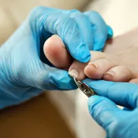a person wearing bright blue disposable gloves is carefully trimming the toenail of another person's second toe. They are using a pair of metal nail nippers to perform the task with precision. The focus is sharp on the hands, the tool, and the toe being attended to, while the background is softly blurred, emphasizing the detailed work being done. The overall impression is one of a clean, hygienic, and professional setting.