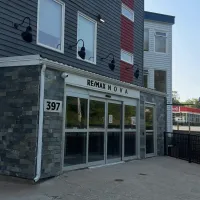 This photograph captures the modern street-front entrance of the RE/MAX NOVA office at 397 bedford highway. The building showcases a contemporary design, combining a dark, stacked-stone facade on the ground level with charcoal siding and a striking red vertical accent on the story above. The entrance features large glass sliding doors under modern black gooseneck lighting. In the background, a Irving gas station can be seen next door.