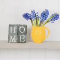On the left, four grey, rustic-style wooden blocks are stacked to spell the word "HOME" in white capital letters. Next to them, a bright yellow ceramic pitcher holds a bouquet of fresh, blue hyacinth flowers.