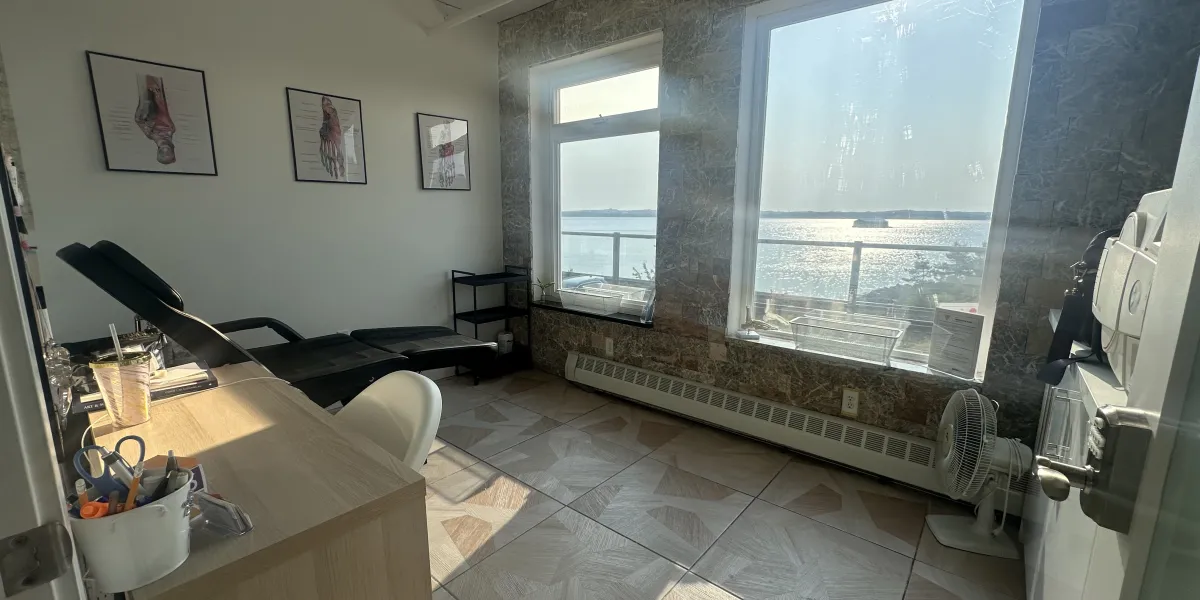 A bright and modern foot care clinic room featuring a black treatment chair, anatomical foot diagrams on the wall, and a wooden desk with office supplies. Large windows along a stone-accented wall provide a scenic view of sparkling water and a clear sky, filling the space with natural light and a tranquil atmosphere.