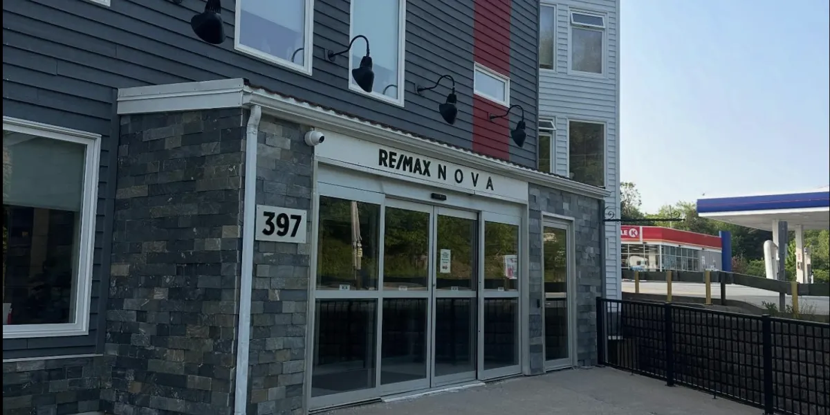 This photograph captures the modern street-front entrance of the RE/MAX NOVA office at 397 bedford highway. The building showcases a contemporary design, combining a dark, stacked-stone facade on the ground level with charcoal siding and a striking red vertical accent on the story above. The entrance features large glass sliding doors under modern black gooseneck lighting. In the background, a Irving gas station can be seen next door.